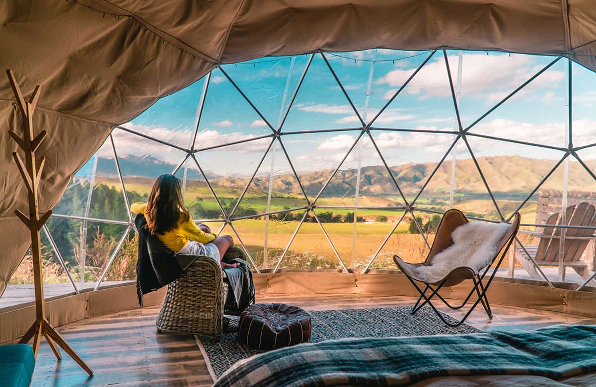 Woman looking out at nature from geo dome tents. Green, blue, orange background. Cozy, camping, glamping, holiday, vacation lifestyle concept. Outdoors cabin, scenic background. New Zealand.