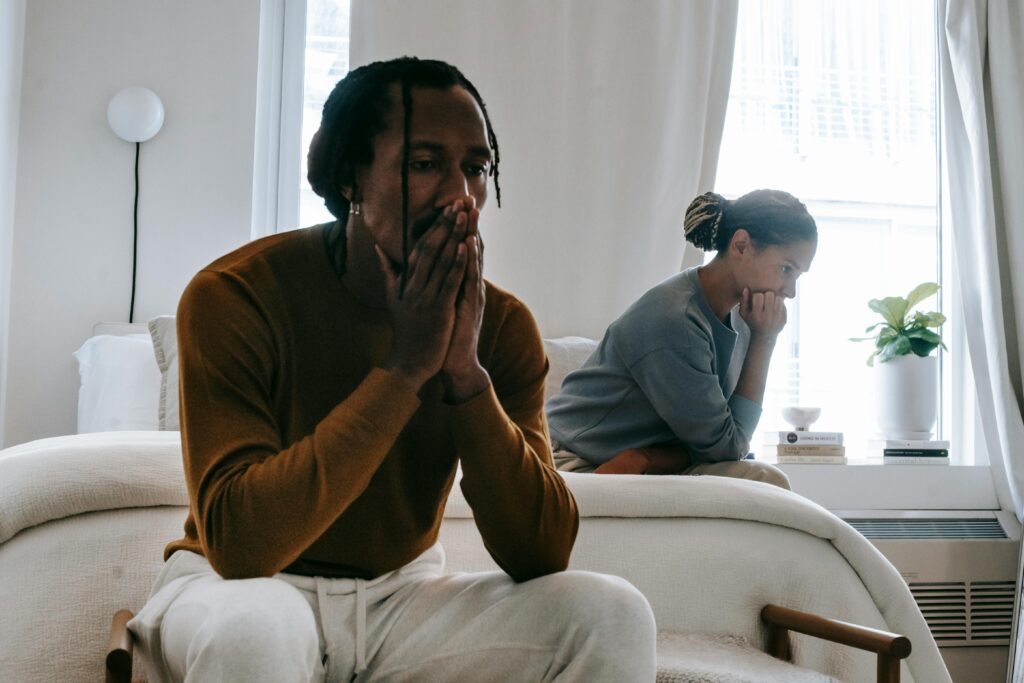 An emotionally distant couple sitting in different parts of the room while one partner looks down at a glowing phone with a soft, intimate expression, suggesting a secretive emotional connection outside the relationship.